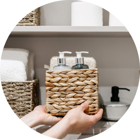 two hands, holding a wicker basket with pump bottles of soap and lotion in it. Folded towels and rolls of toilet paper are in the background.