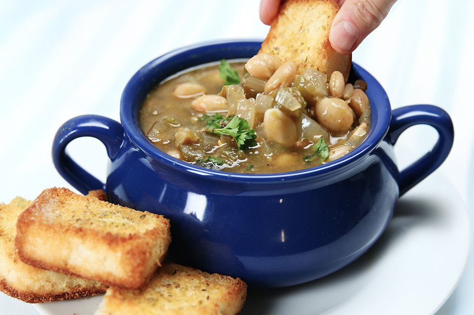 The recipe detailed in this article: a Spicy White Bean chili, served in a blue bowl with handles and some toasted bread on the side. A hand is dipping a piece of bread into the chili.