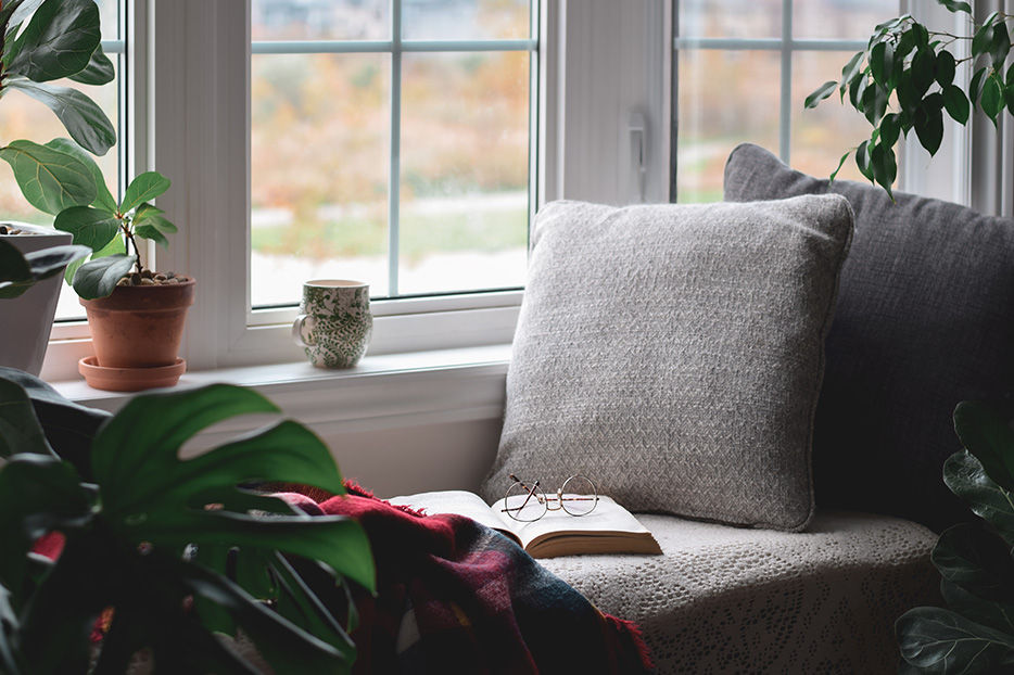 A cozy spot by the window with an opened book and glasses sitting on it. Pillows, a blanket, and a coffee mug make it look particularly cozy